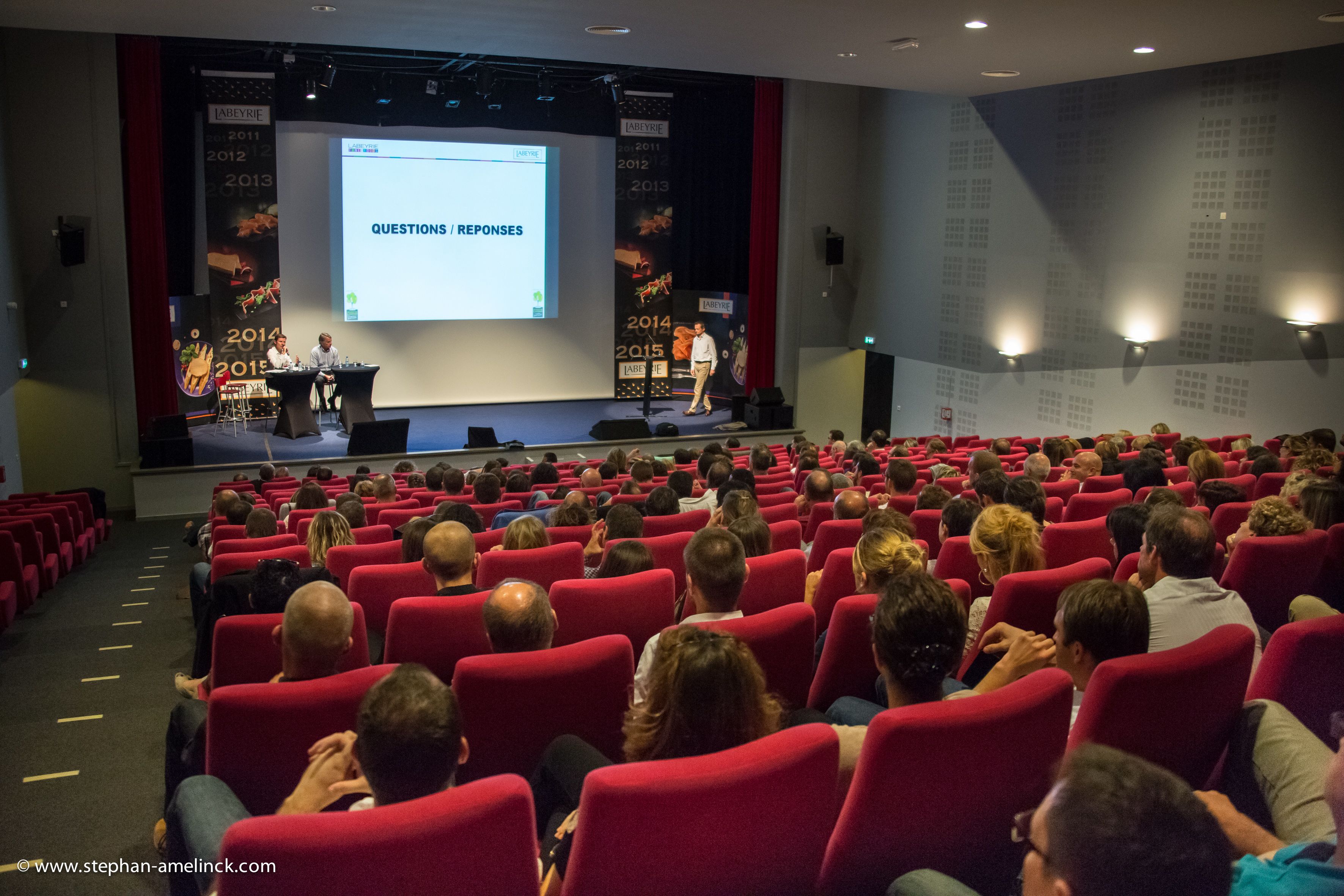 Uma sala de conferências com fileiras de cadeiras vermelhas voltadas para um palco com uma tela de apresentação.