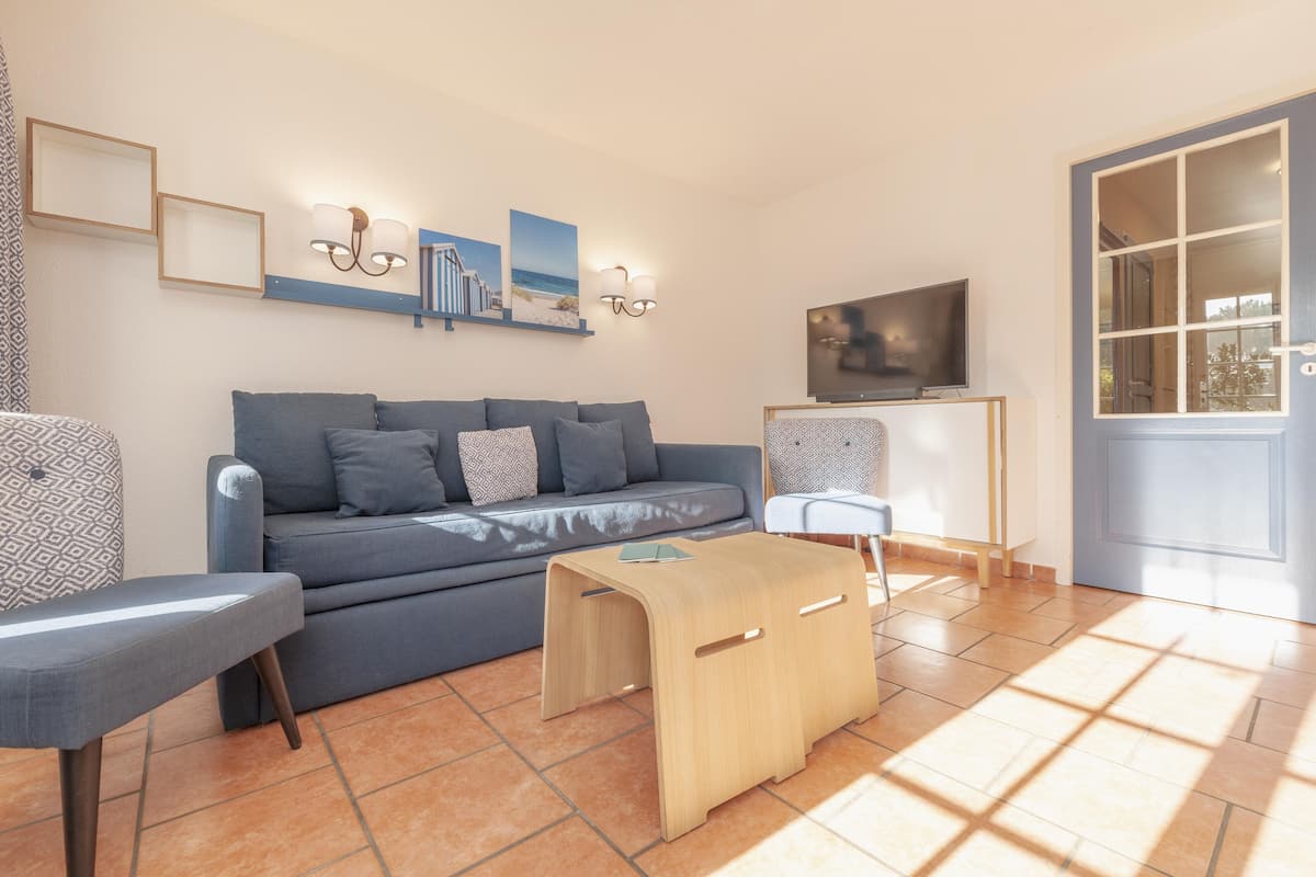 Una sala de estar moderna con un sofá gris, una mesa de centro de madera y un televisor sobre un mueble. Hay luces fijadas en la pared e imágenes enmarcadas sobre el sofá.