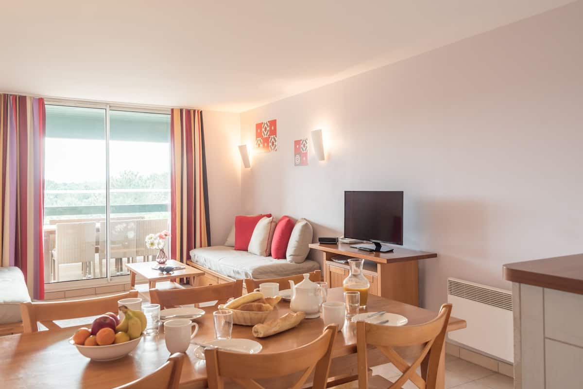 Una habitación moderna con una mesa de comedor lista para el desayuno, un sofá con almohadones rojos, un televisor y un balcón con vistas.
