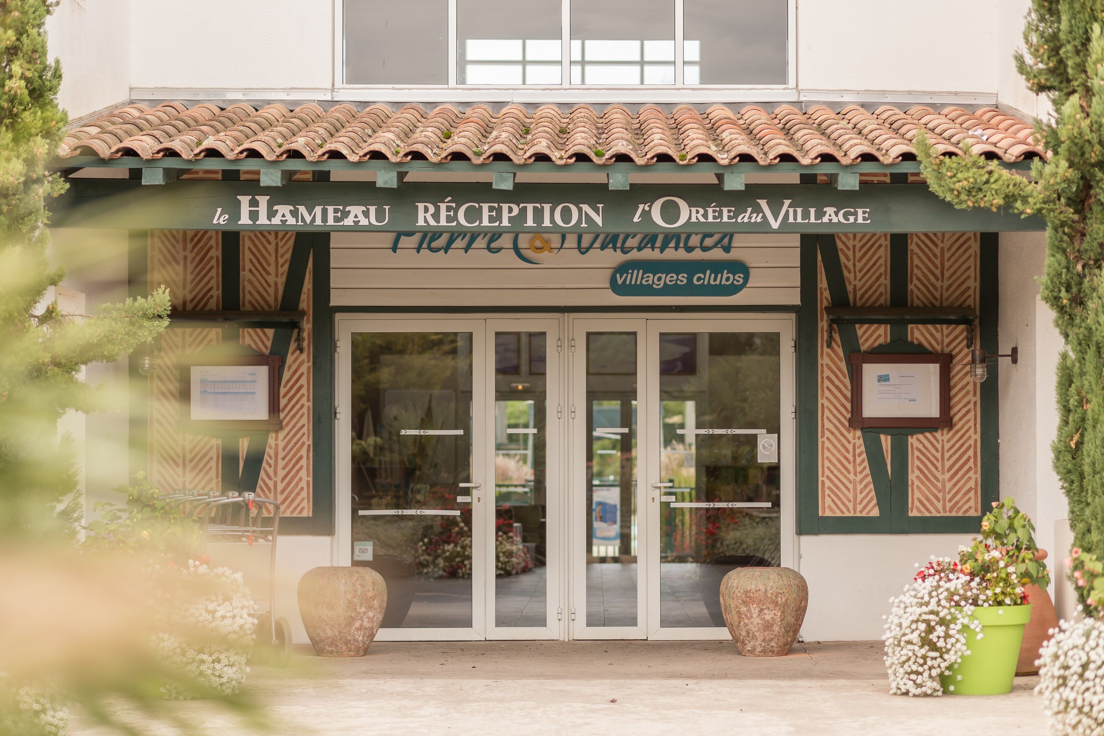 L’entrée d’un hôtel, avec un toit en tuiles, des portes en verre et une enseigne indiquant : « Le Hameau, réception de l’Orée du Village. »