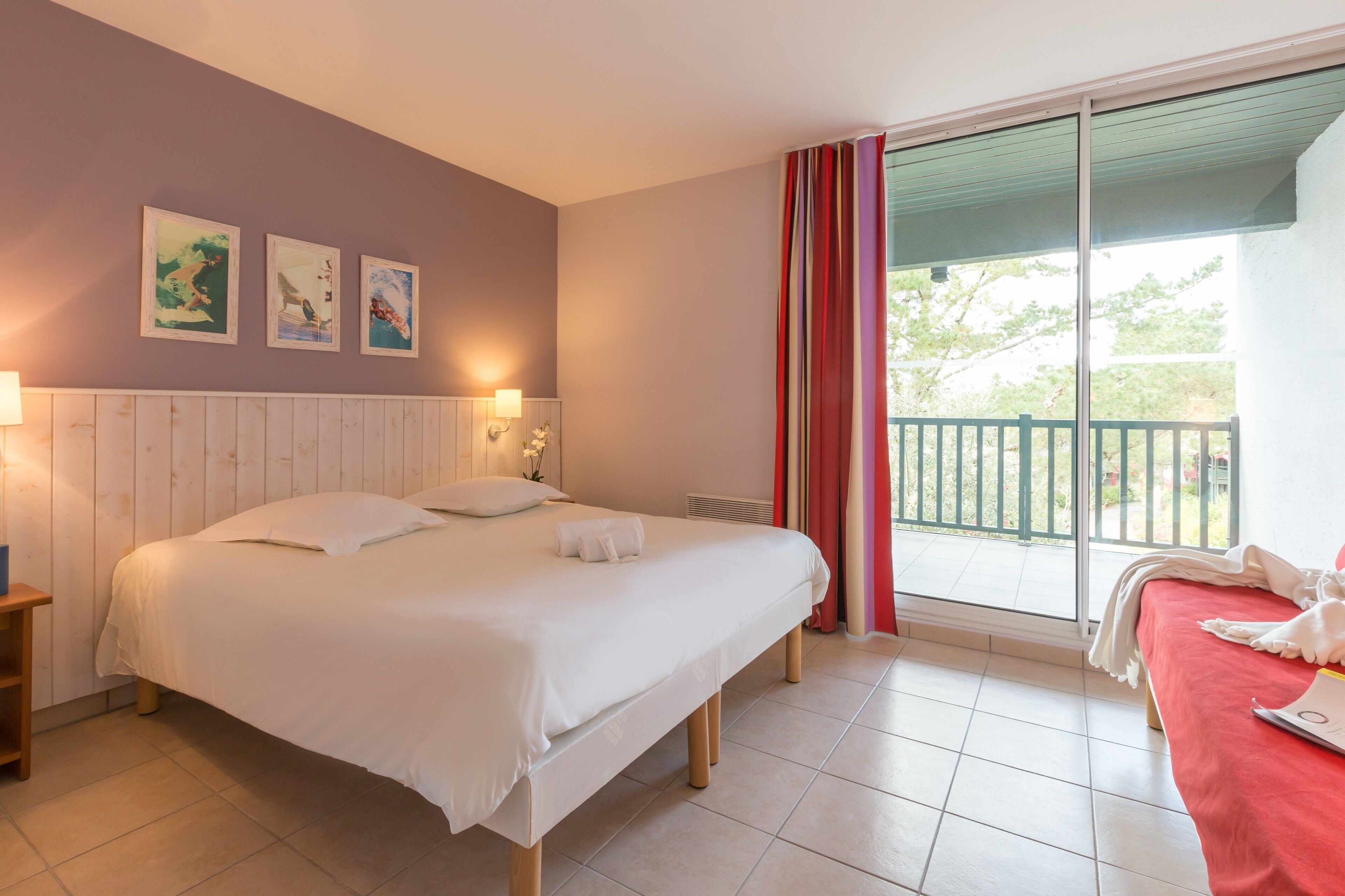 Una camera d'albergo con un grande letto, un comodino, una finestra con tende rosse e un quadro appeso al muro.