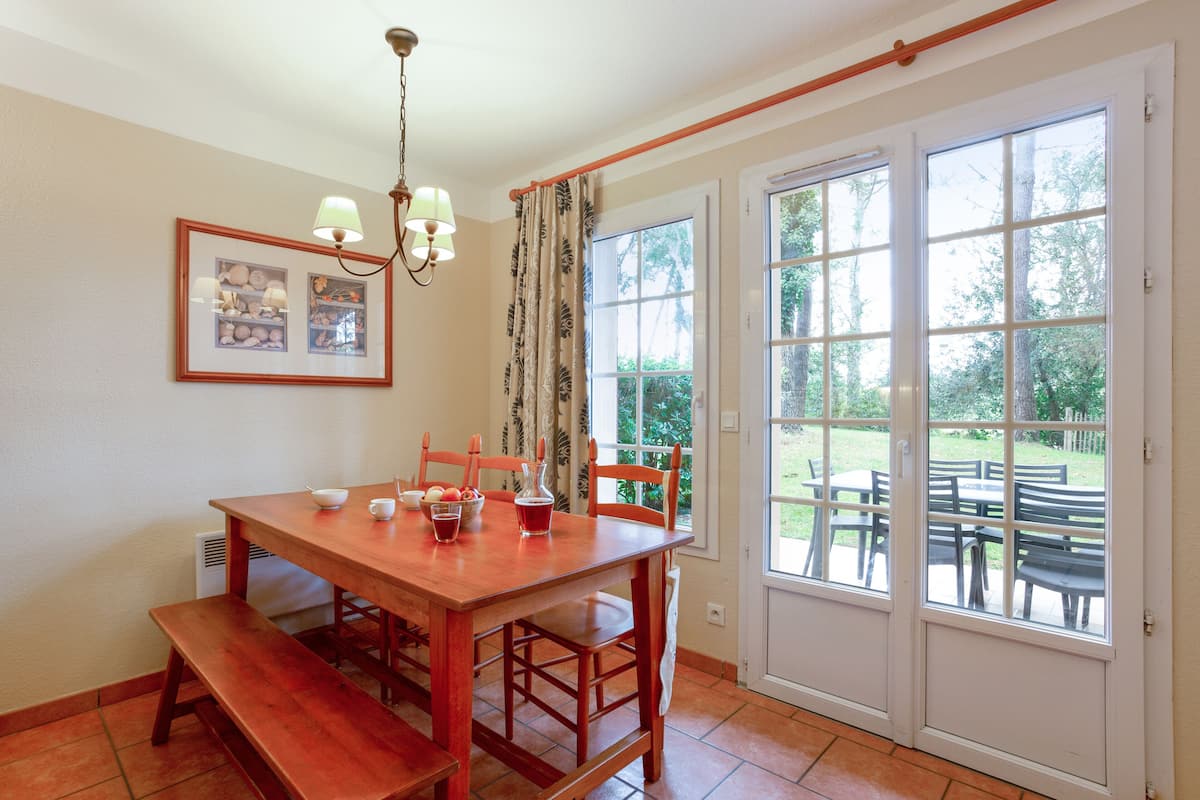 Un comedor con mesa de madera, sillas, una banca, una araña y un ventanal con vistas al exterior.