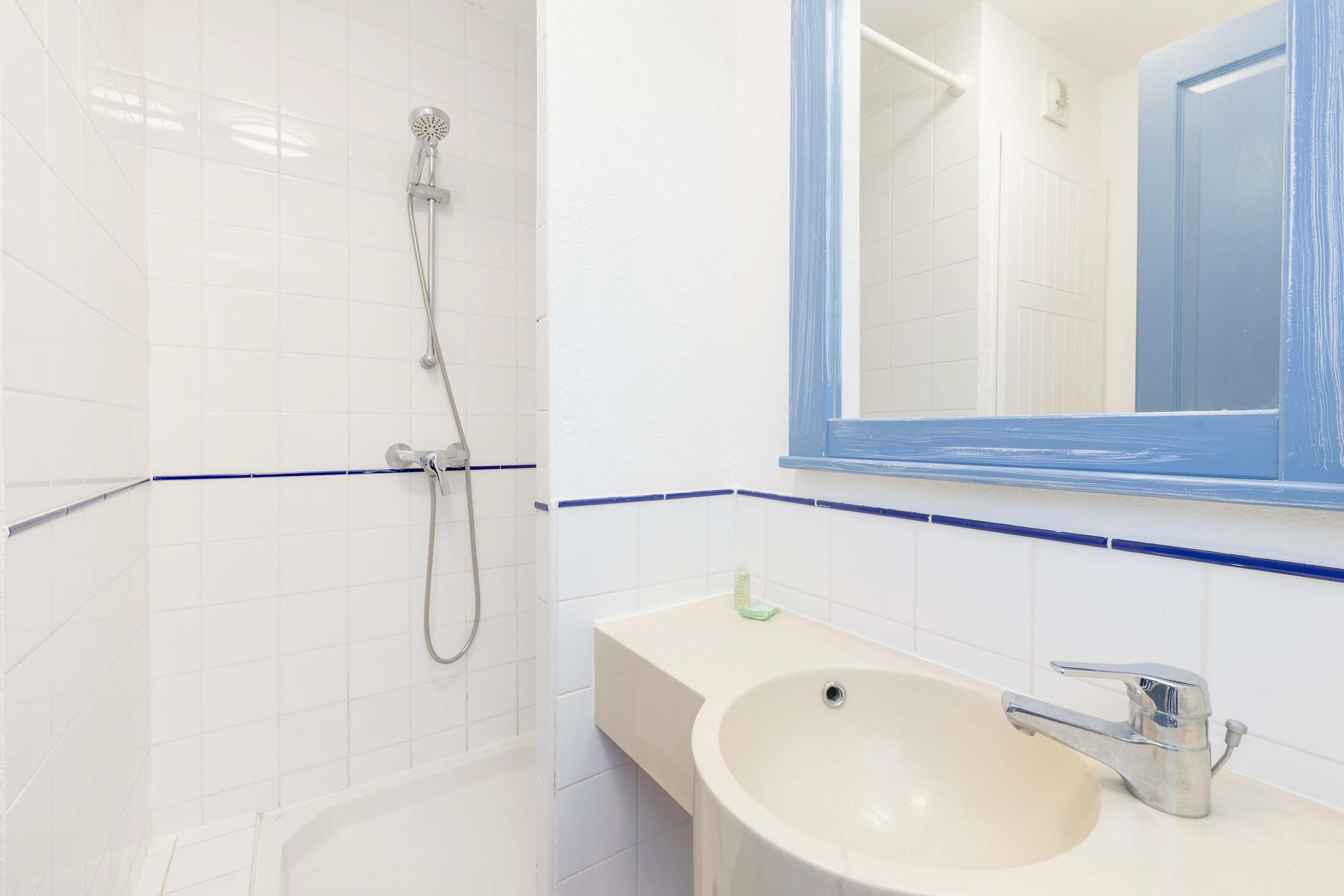 Un baño con paredes de azulejos blancos, un lavamanos color beige, un espejo con marco azul y una ducha con cabezal de ducha de mano.