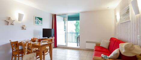 Ein Zimmer mit Esstisch, Stühlen, Fernseher, einem roten Sofa und einem Balkon mit rotem Vorhang.