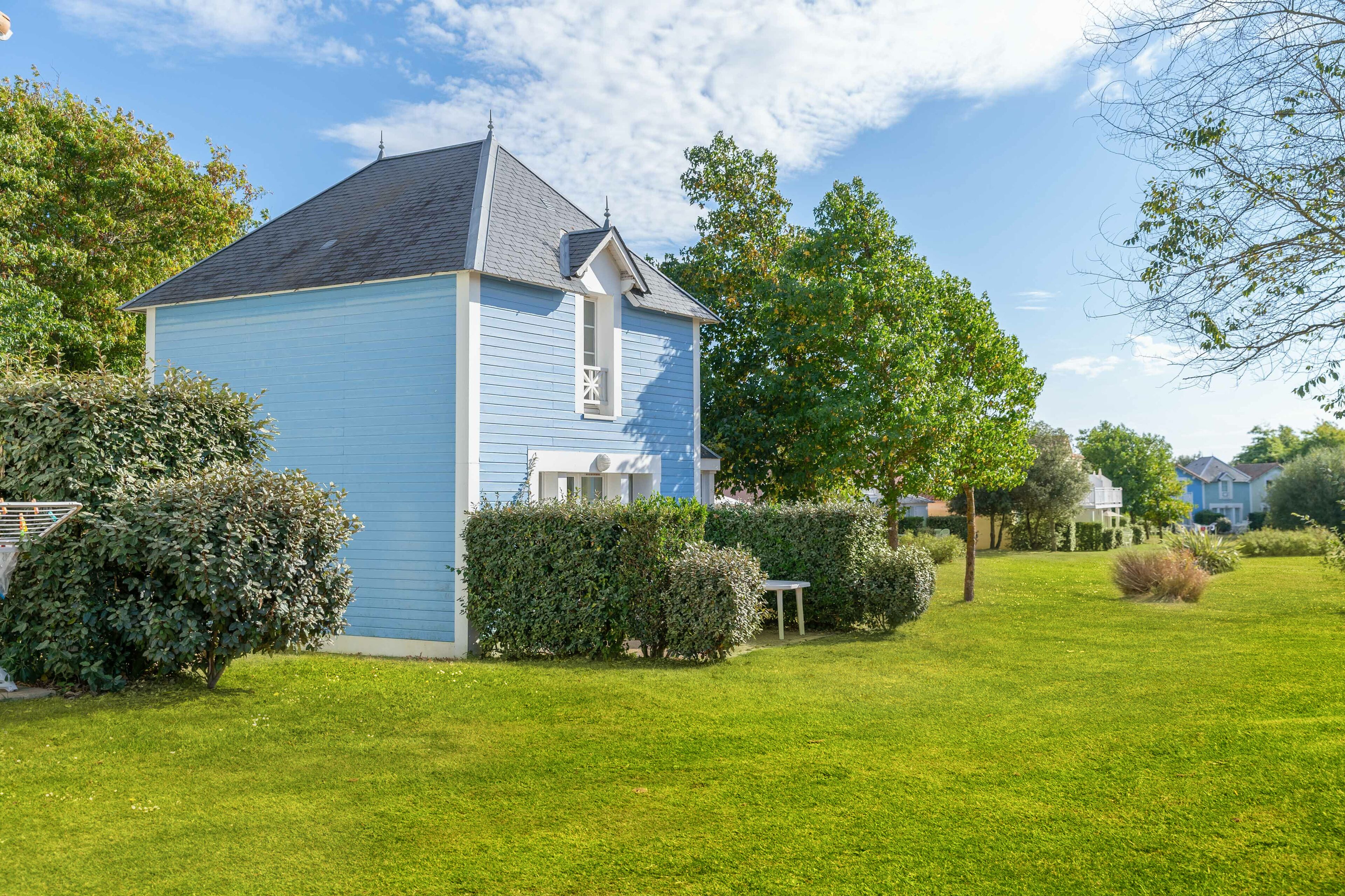 A small, light blue house with a gabled roof, surrounded by a well-maintained lawn and various shrubs.