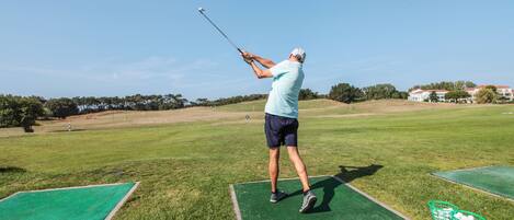 Un campo de golf con cielo despejado, un golfista en pleno balanceo y canastas verdes sobre el césped.