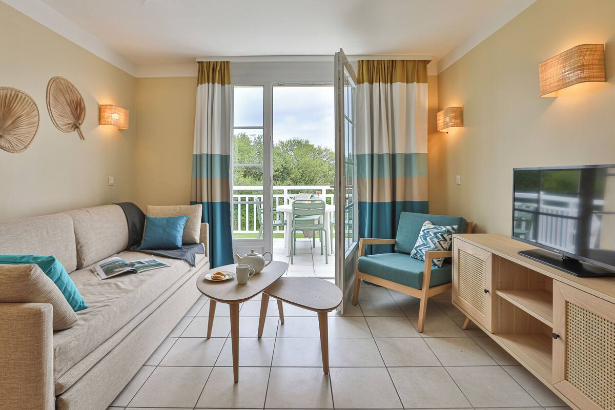 Een moderne woonkamer met een bank, een klein tafeltje, een televisie en uitzicht op het buitenleven door een glazen deur.