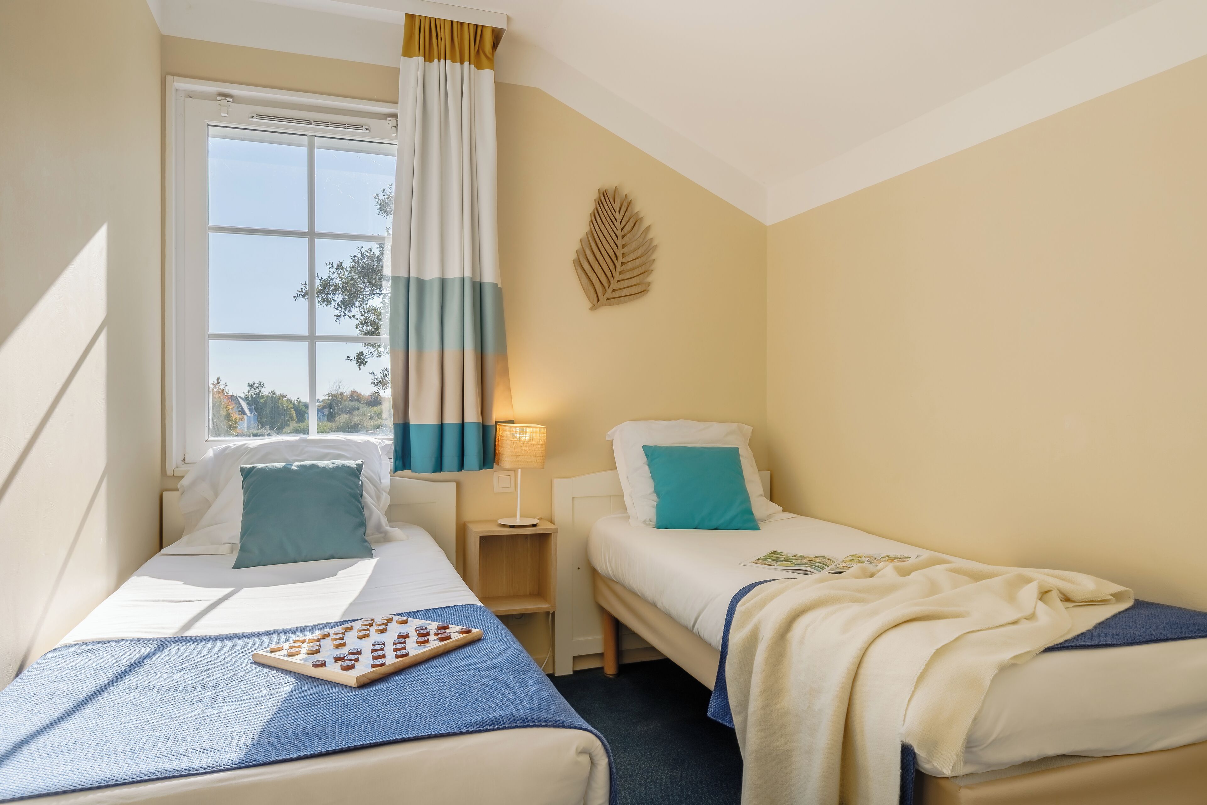 Habitación de hotel con dos camas, una ventana con cortinas, un adorno de hojas en la pared y una mesita de noche con lámpara.