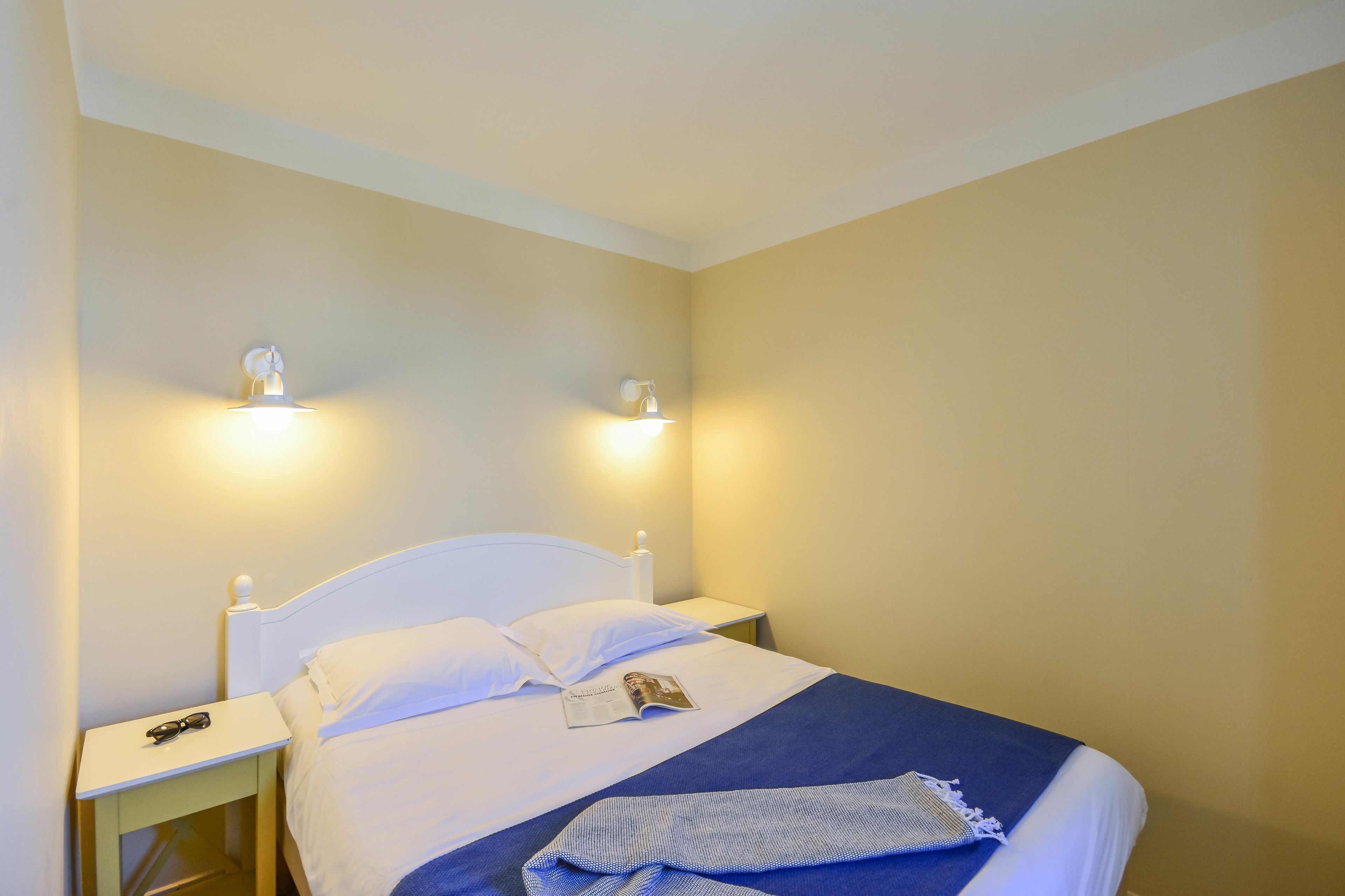 A neatly made bed with white linens and a blue blanket, two bedside lamps, and a magazine on the bed.