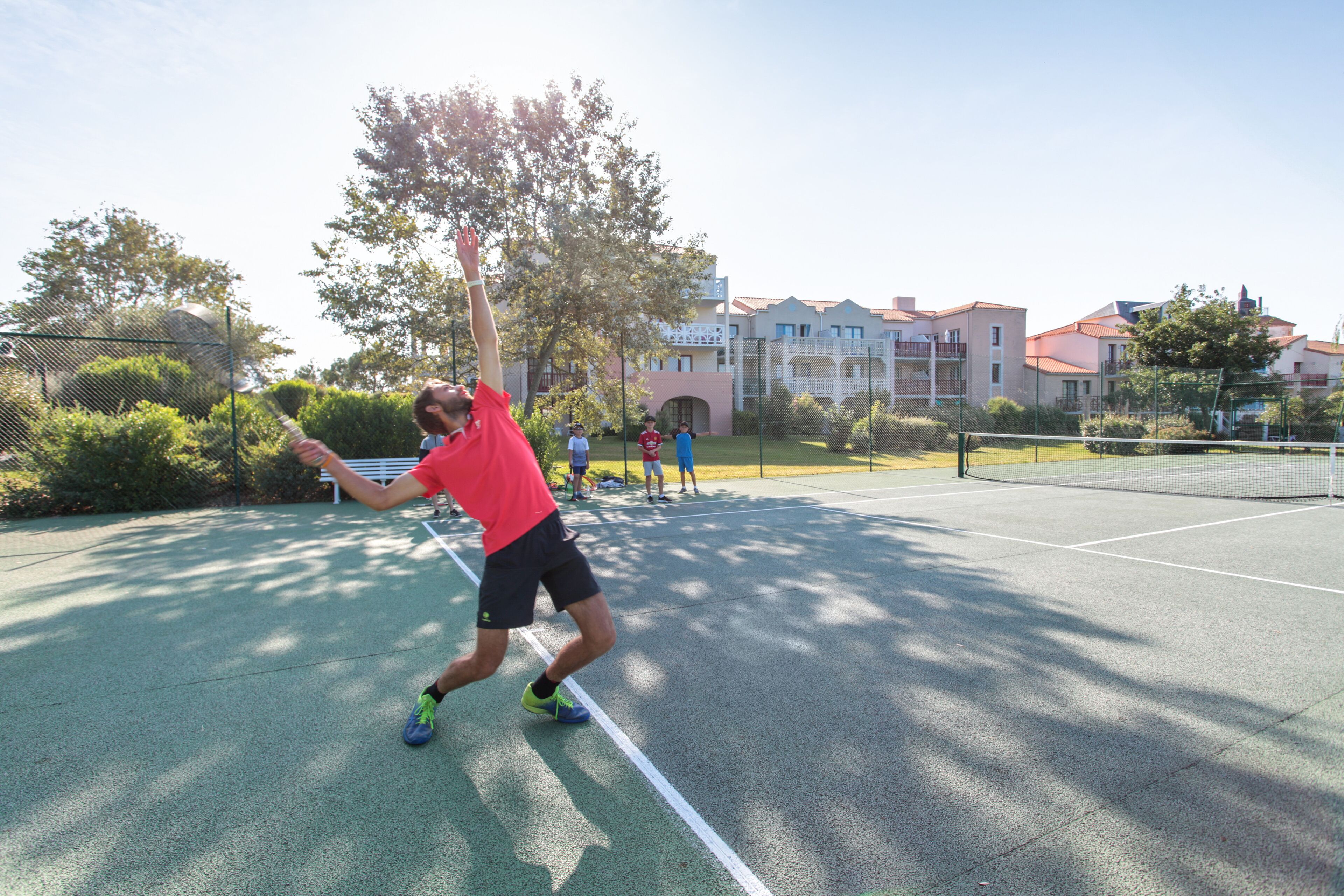 Een tennisbaan met een net, omringd door groen en gebouwen.