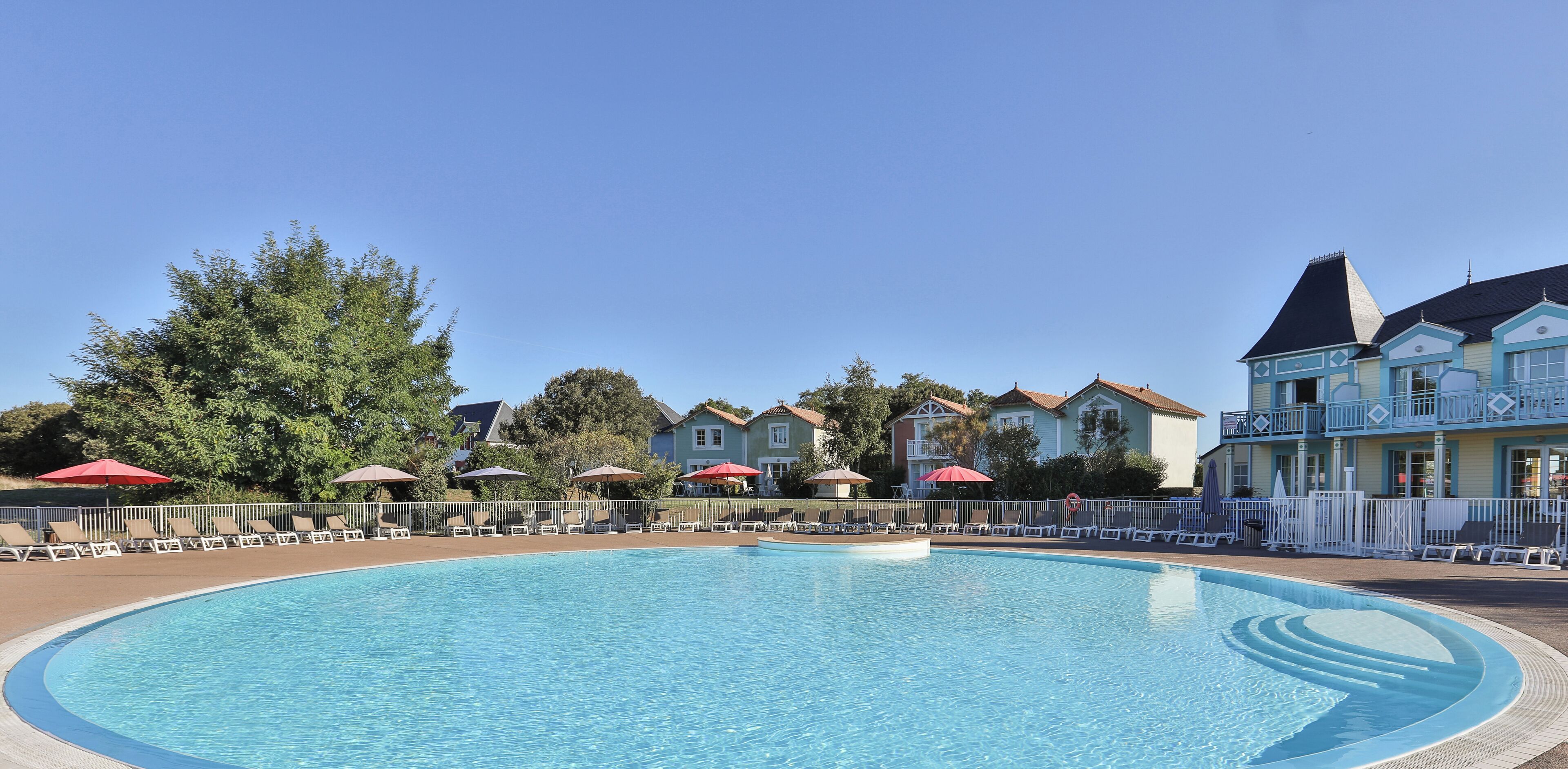 A swimming pool with sun loungers and umbrellas, surrounded by houses with balconies.