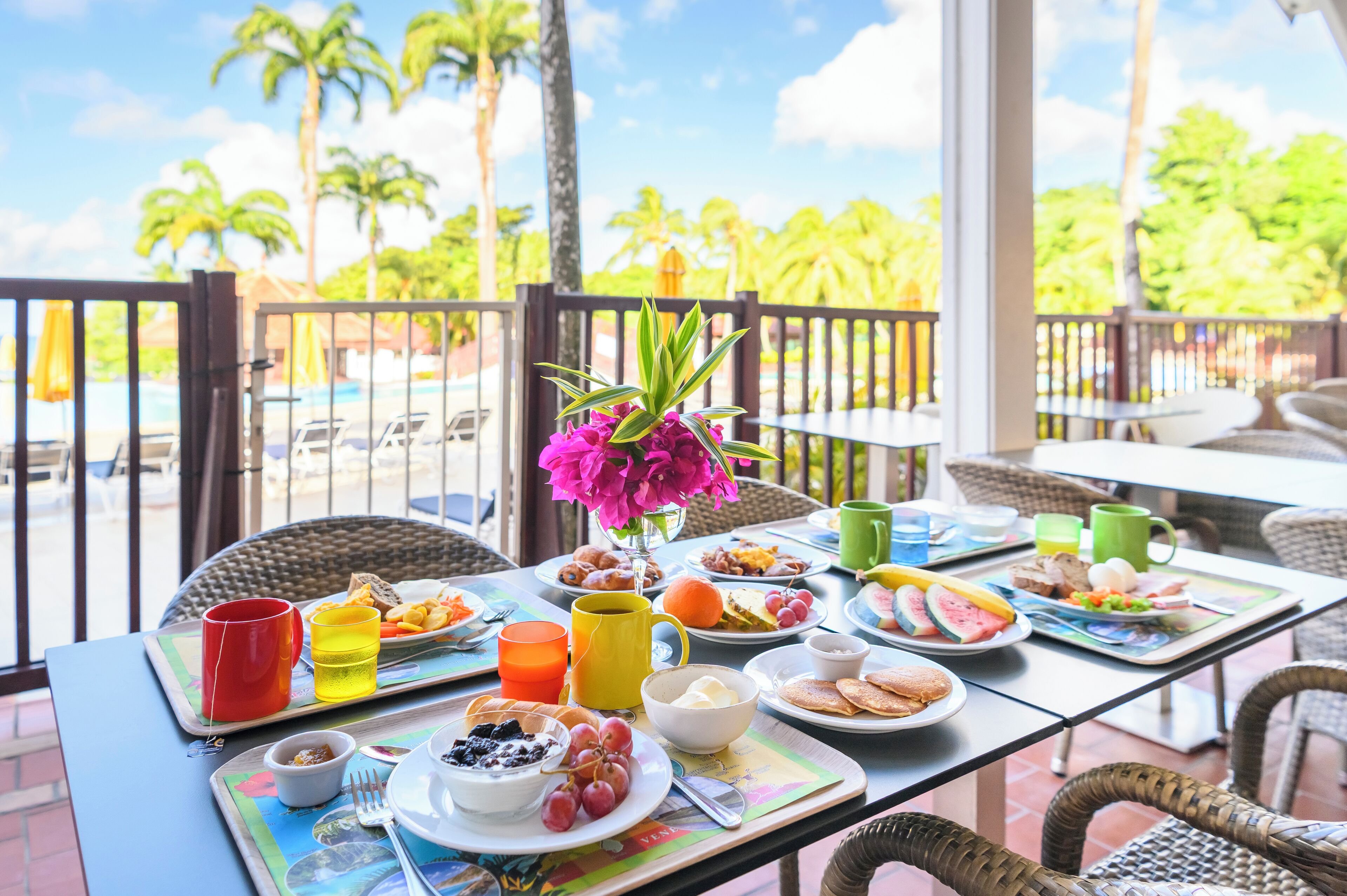 A breakfast table with various dishes, drinks, and a vase of flowers, set on a balcony with a view of palm trees and a pool.