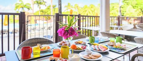 A breakfast table with various dishes, drinks, and a vase of flowers, set on a balcony with a view of palm trees and a pool.