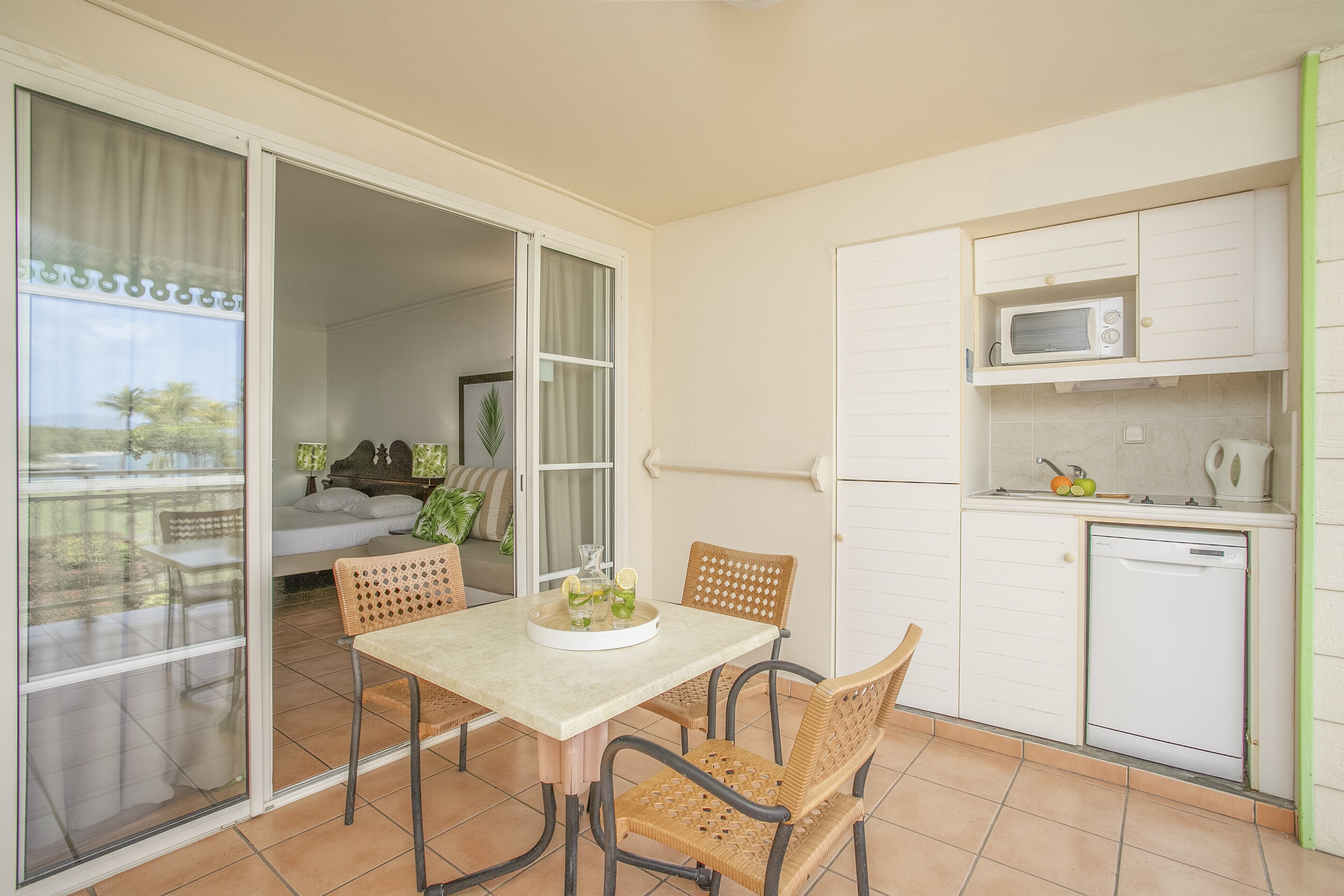 A compact kitchen with a microwave and dishwasher, a dining area with wicker chairs, and a sliding glass door leading to a balcony.