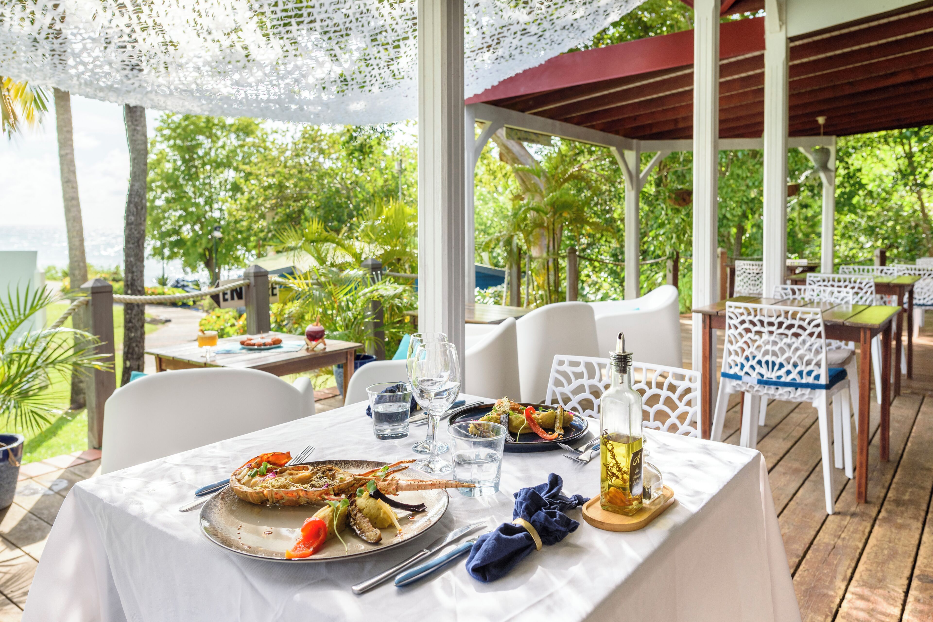 Un espace de restauration en plein air avec une table dressée pour deux personnes, entouré de chaises blanches, un coin salon abrité et une végétation luxuriante en arrière-plan.