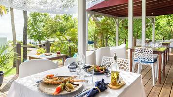 Un espace de restauration en plein air avec une table dressée pour deux personnes, entouré de chaises blanches, un coin salon abrité et une végétation luxuriante en arrière-plan.