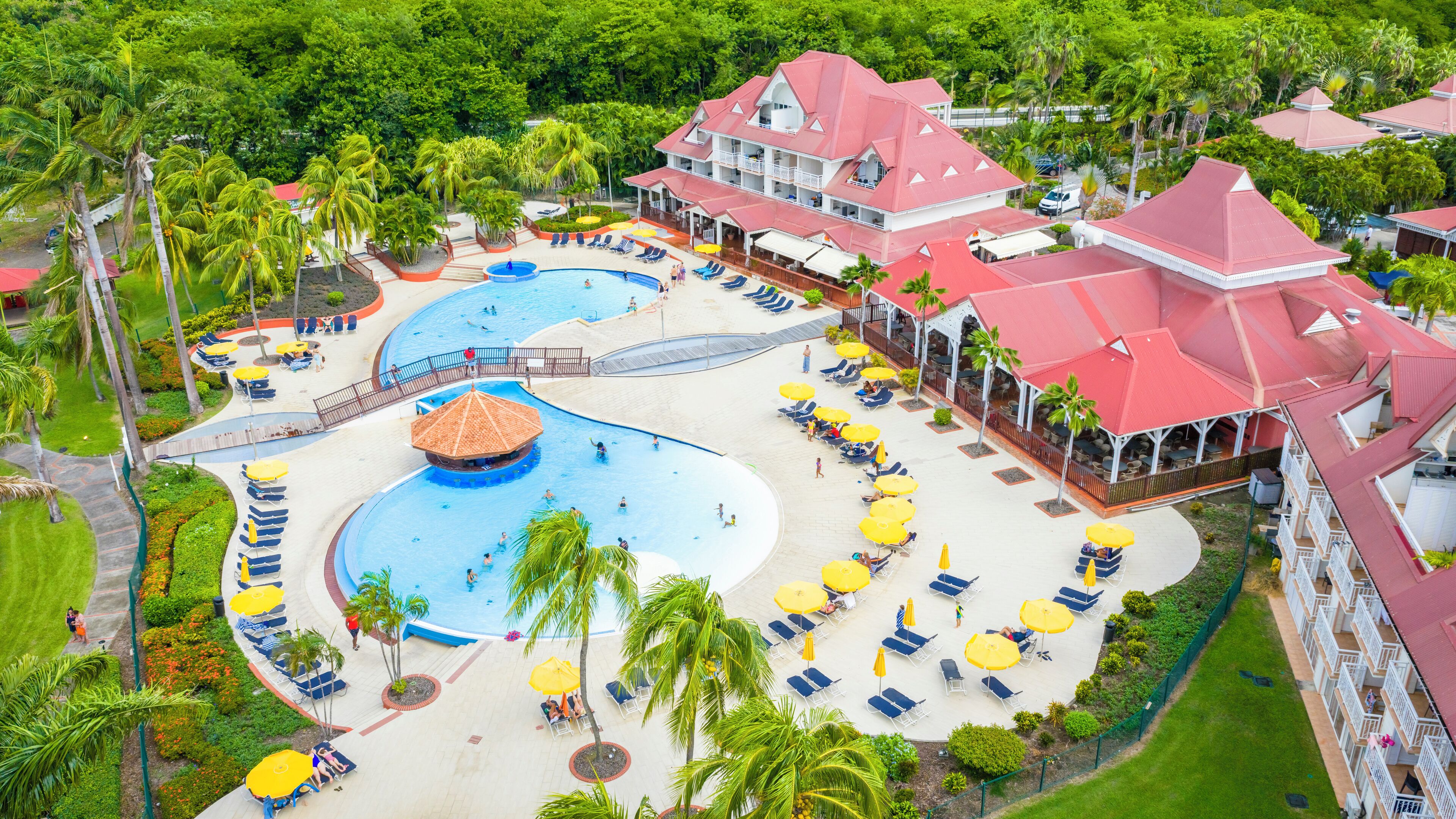 Un resort con una amplia zona de piscina, varias áreas de descanso con sombrillas y un edificio destacado con tejado rojo.