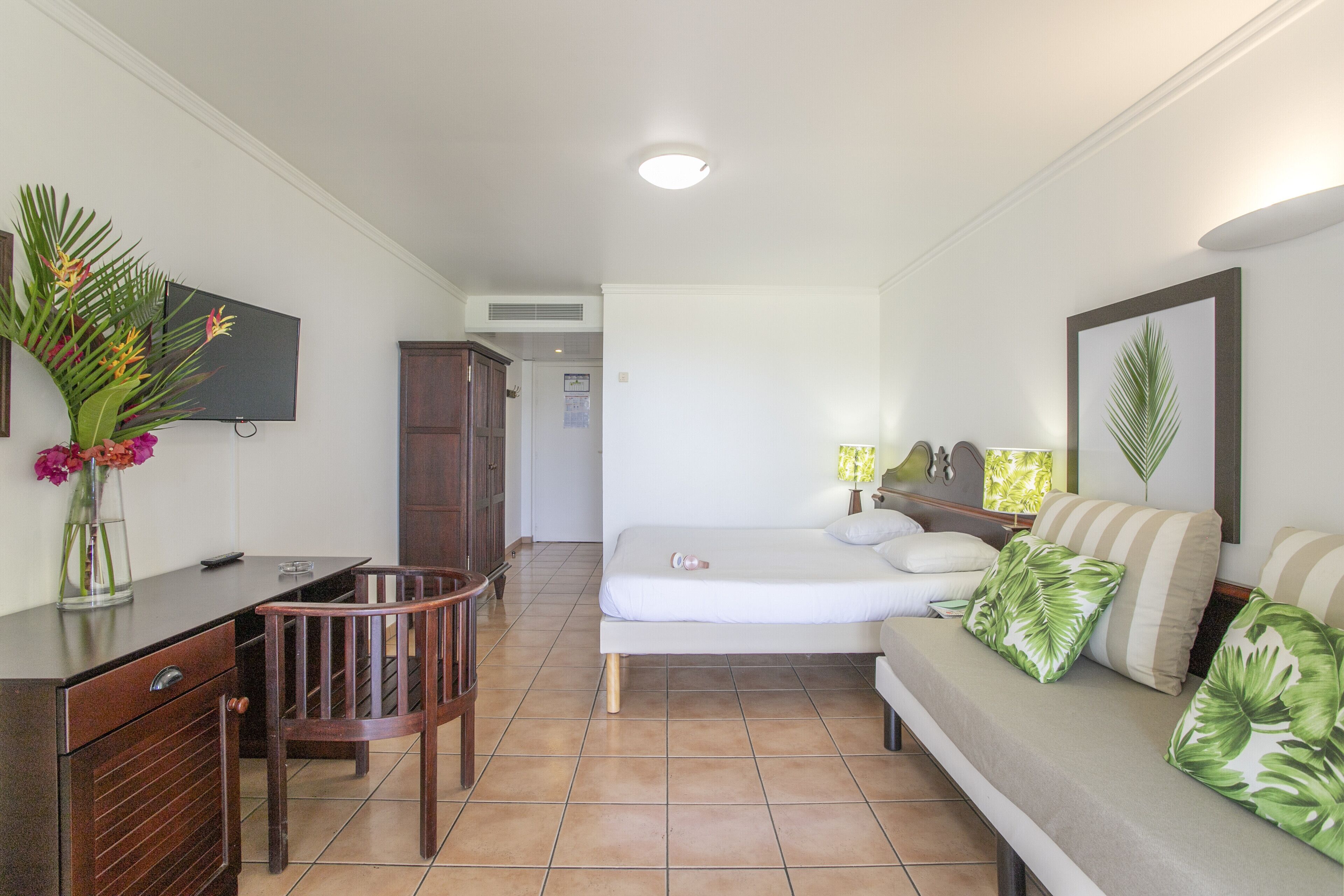 Habitación de hotel con cama, sofá, escritorio con silla, televisor montado en la pared y un jarrón con flores sobre el escritorio.