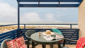 Un balcon donnant sur la plage, avec une table sur laquelle on trouve un chapeau, des lunettes de soleil et un sac à rayures.