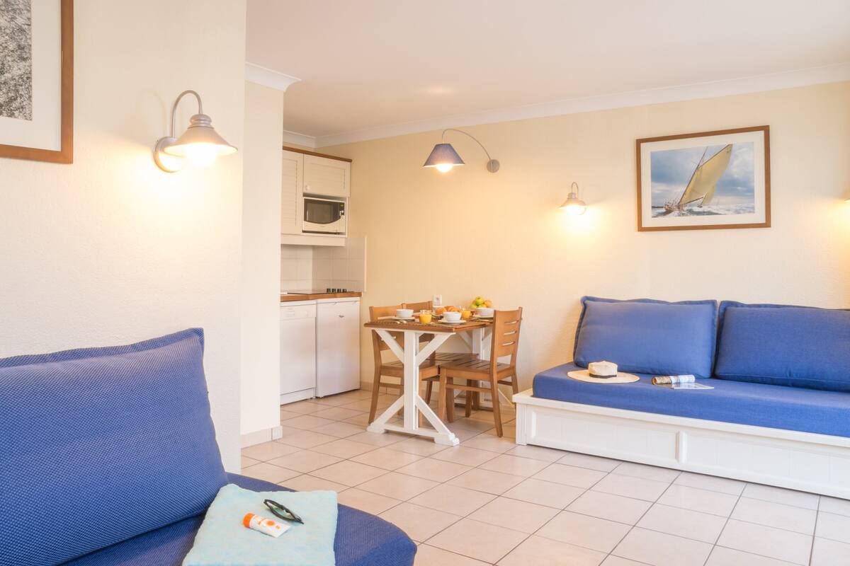 Un salon avec un canapé bleu, une table à manger avec des chaises, un coin cuisine et un tableau encadré au mur.