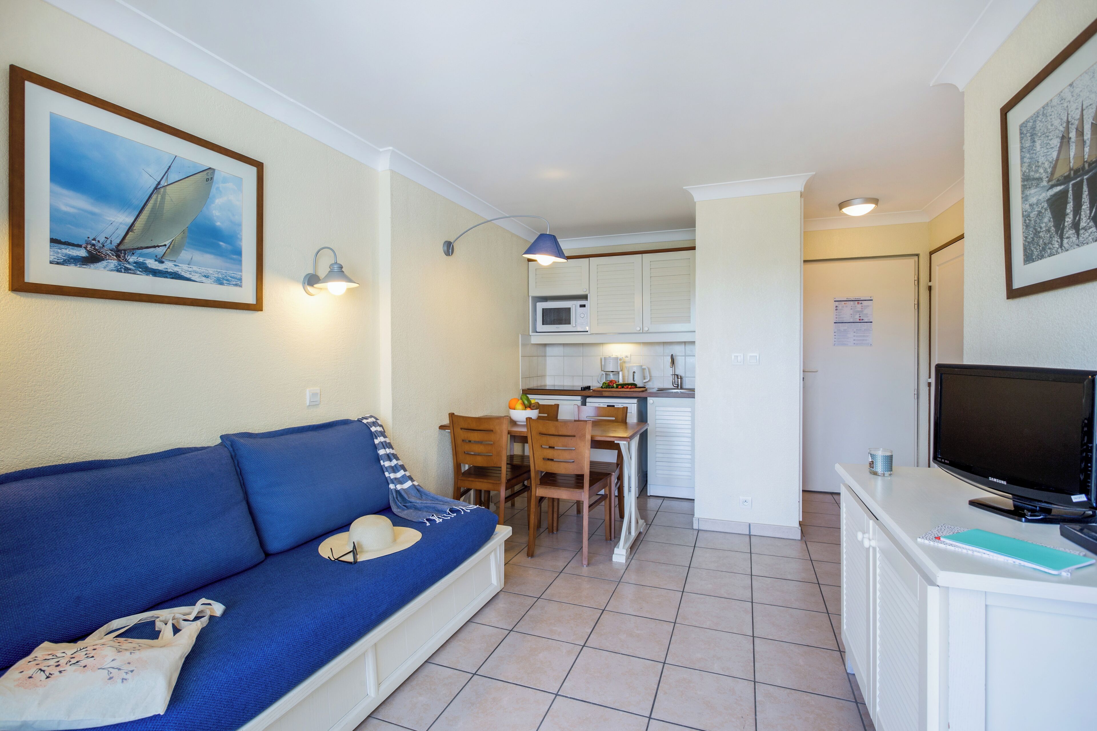 Un espacio habitable compacto con un sofá azul, una zona de comedor, una cocina con microondas y un televisor sobre un mueble blanco.