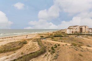 Uma área costeira com uma praia arenosa, um caminho para pedestres e um complexo de edifícios com vários andares e varandas com vista para a praia.