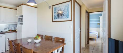A compact kitchen with white cabinets, a dining table with chairs, a fruit bowl, and a framed picture on the wall.
