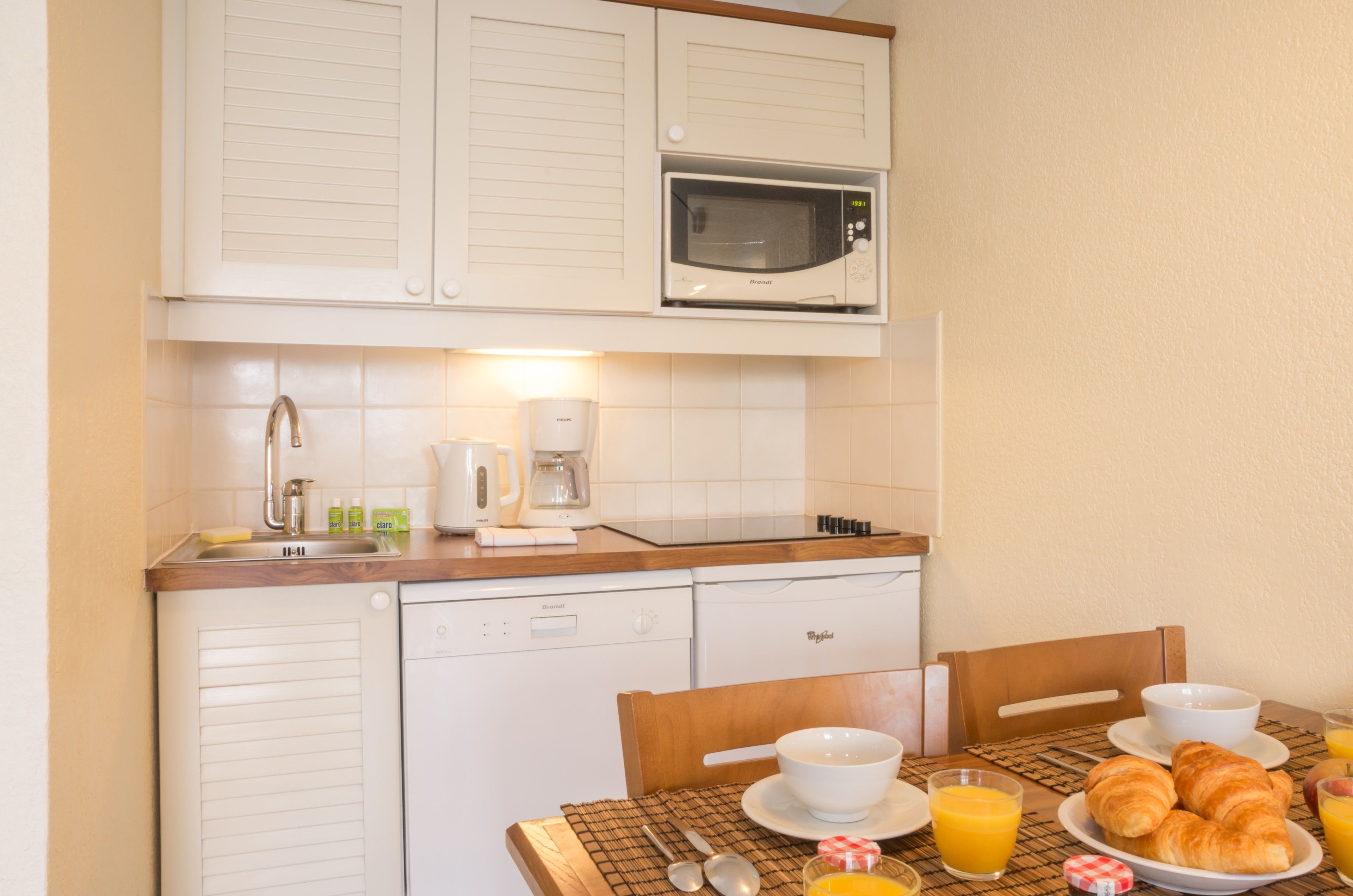A compact kitchen with a microwave, coffee maker, and dishwasher. There is a breakfast table with croissants, juice, and a bowl.