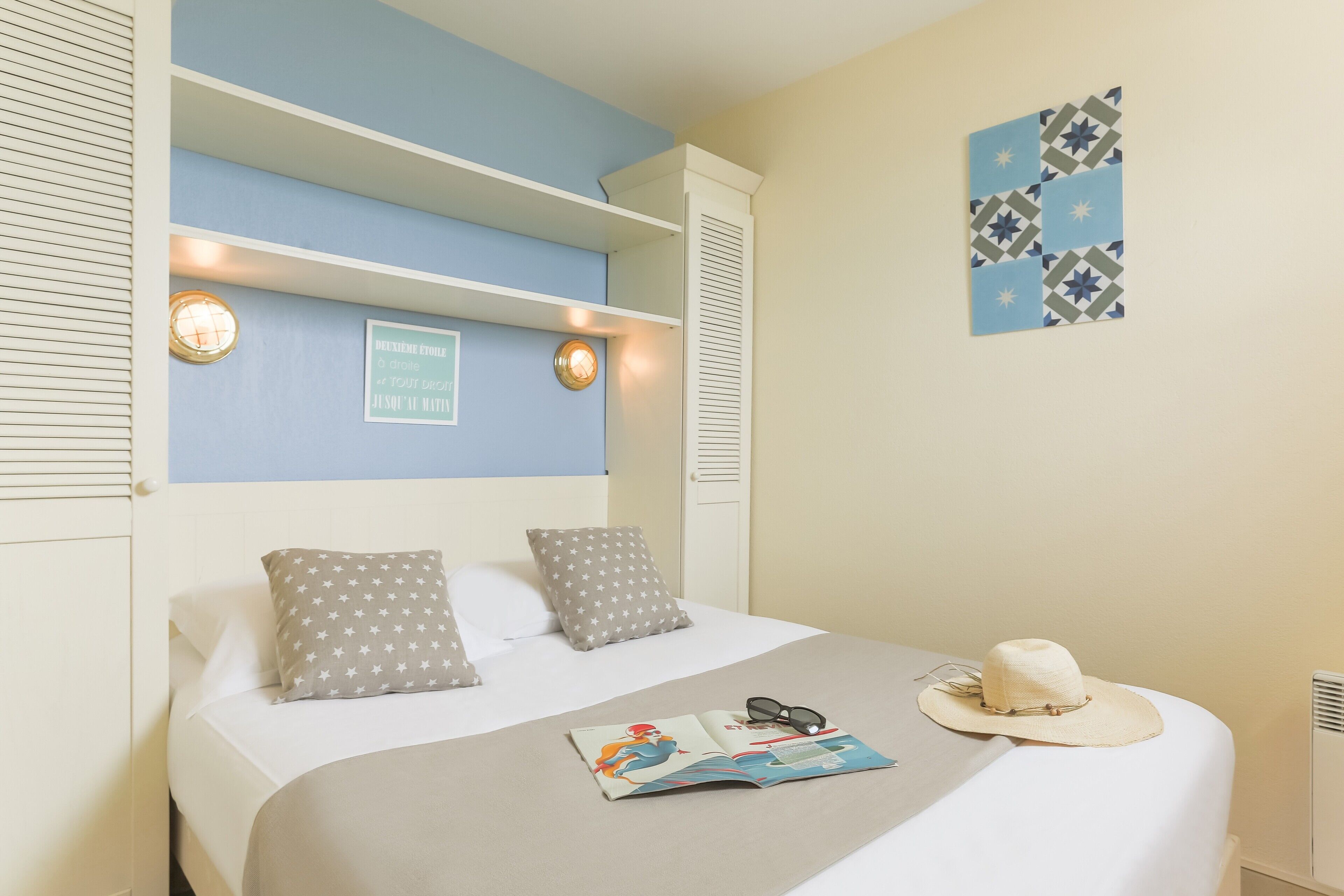 A neatly made bed with star-patterned pillows, a magazine, and a hat on the bed. There are wall sconces and a framed quote on the wall.