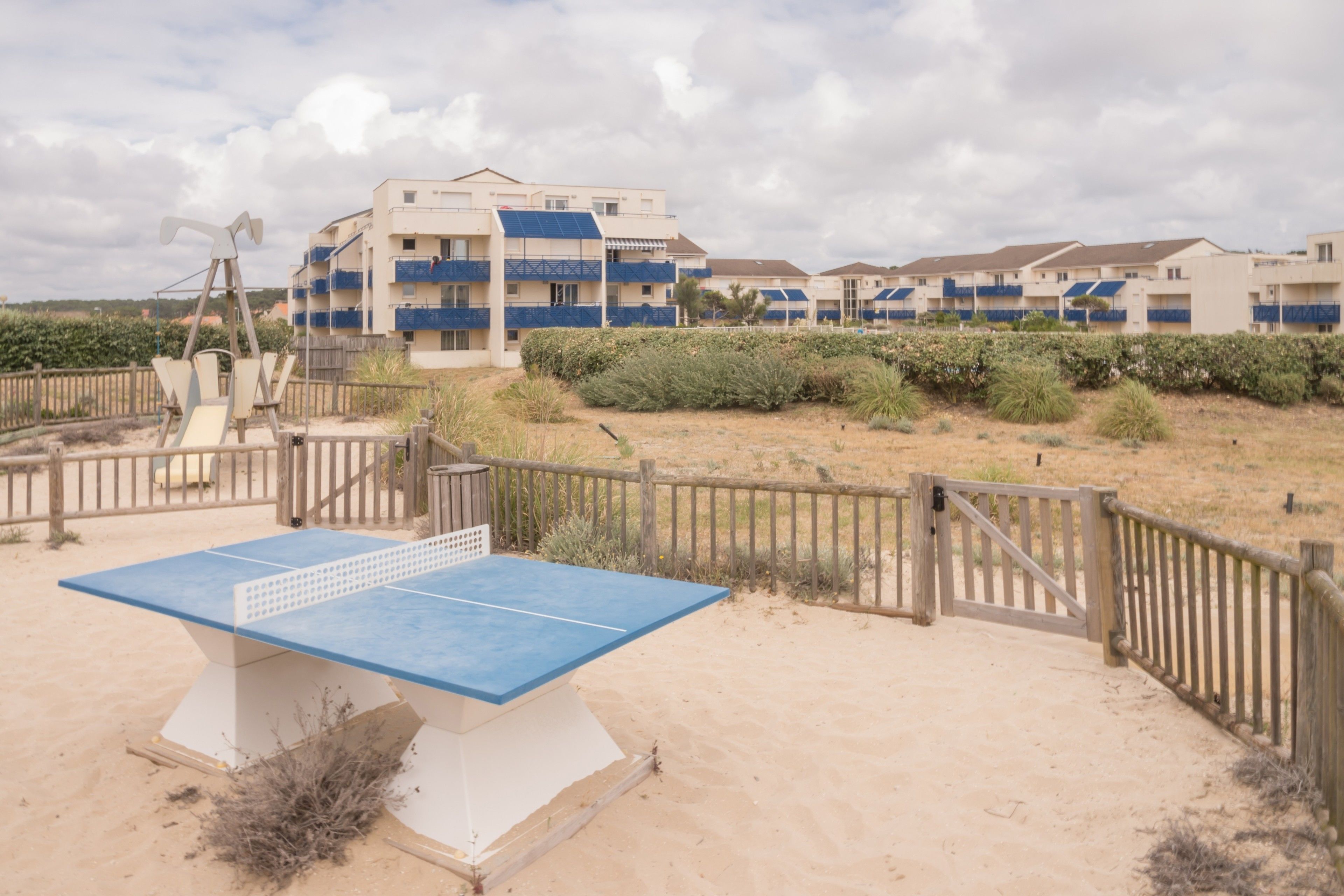 A beachside table tennis court with a wooden fence and a building with blue balconies in the background.