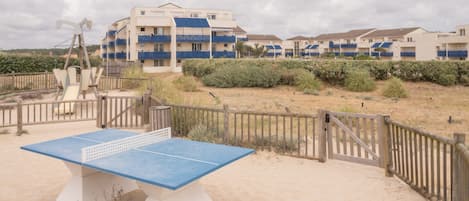 A beachside table tennis court with a wooden fence and a building with blue balconies in the background.