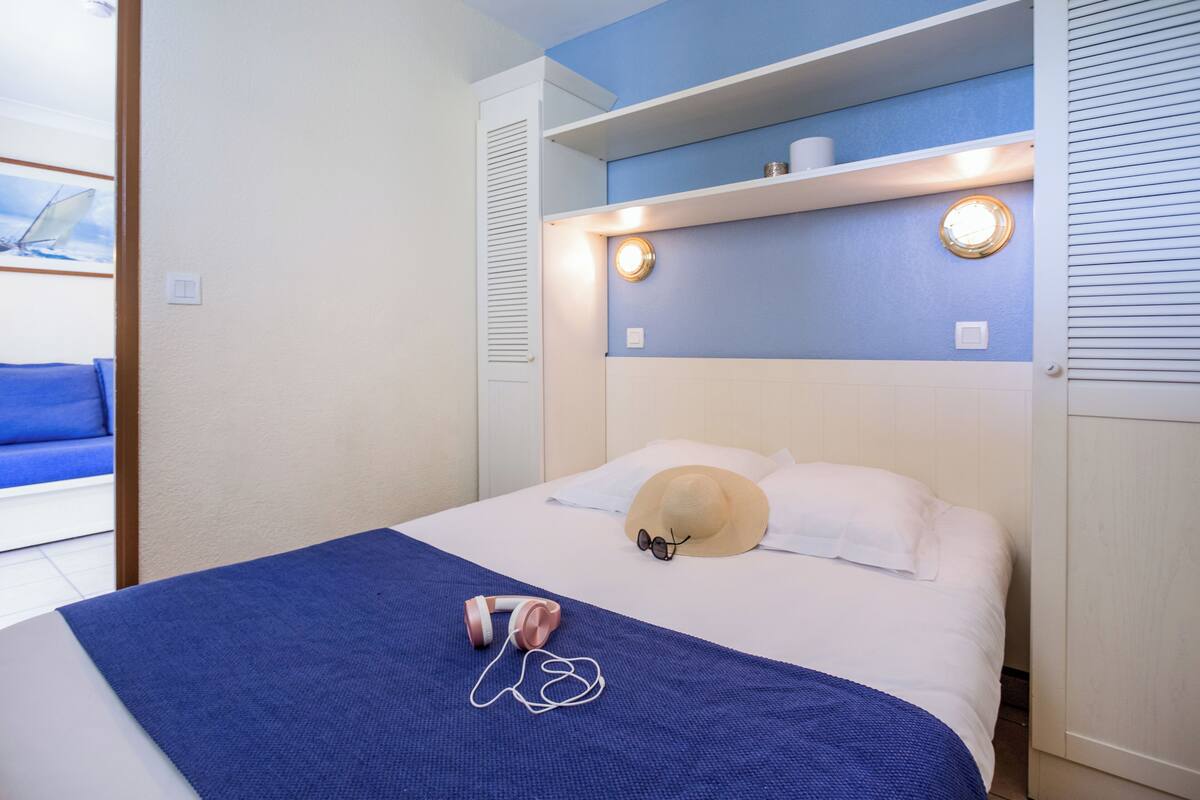 Un lit bien fait, recouvert d’une couverture bleue, avec un chapeau beige et une paire d’écouteurs posés dessus.