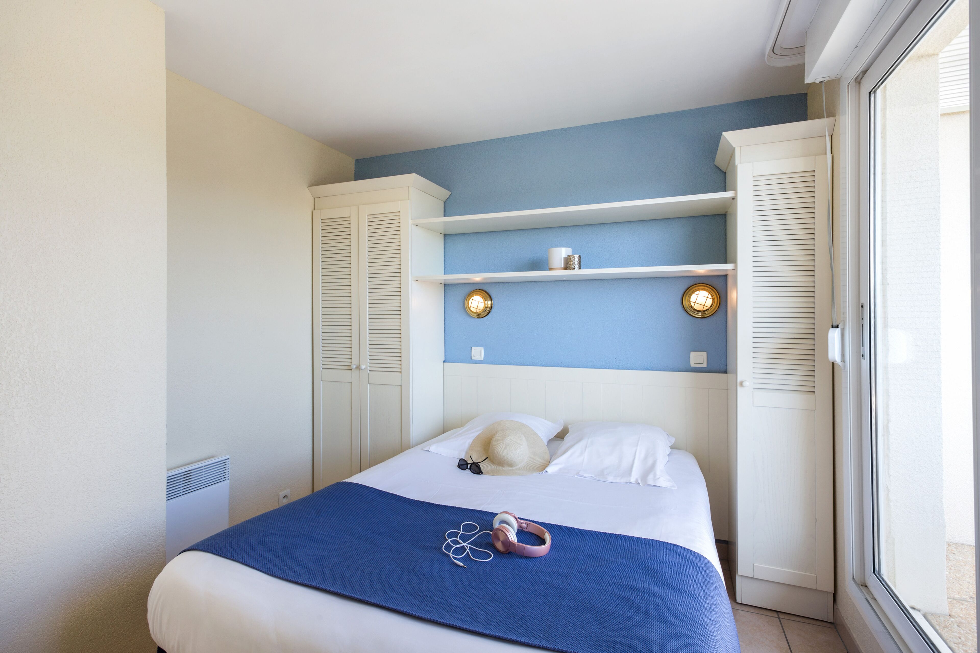 A bedroom with a bed, a blue blanket, a hat, and headphones on the bed. There are white wardrobes and a window with blinds.