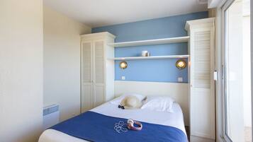 Ein Schlafzimmer mit einem Bett, einer blauen Decke, einem Hut und Kopfhörern auf dem Bett. Weiße Schränke und ein Fenster mit Jalousien sind ebenfalls vorhanden.