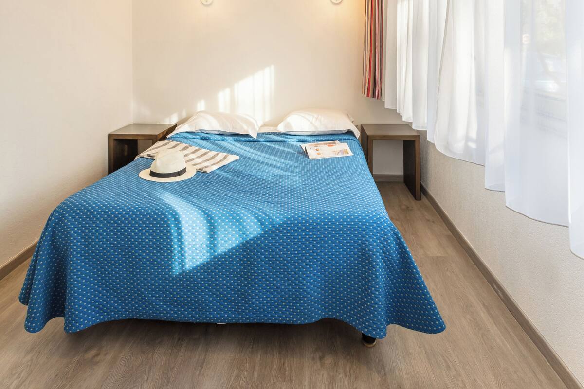 Una habitación de hotel con una cama, dos mesitas de noche, dos lámparas de pared y un ventanal con cortinas.