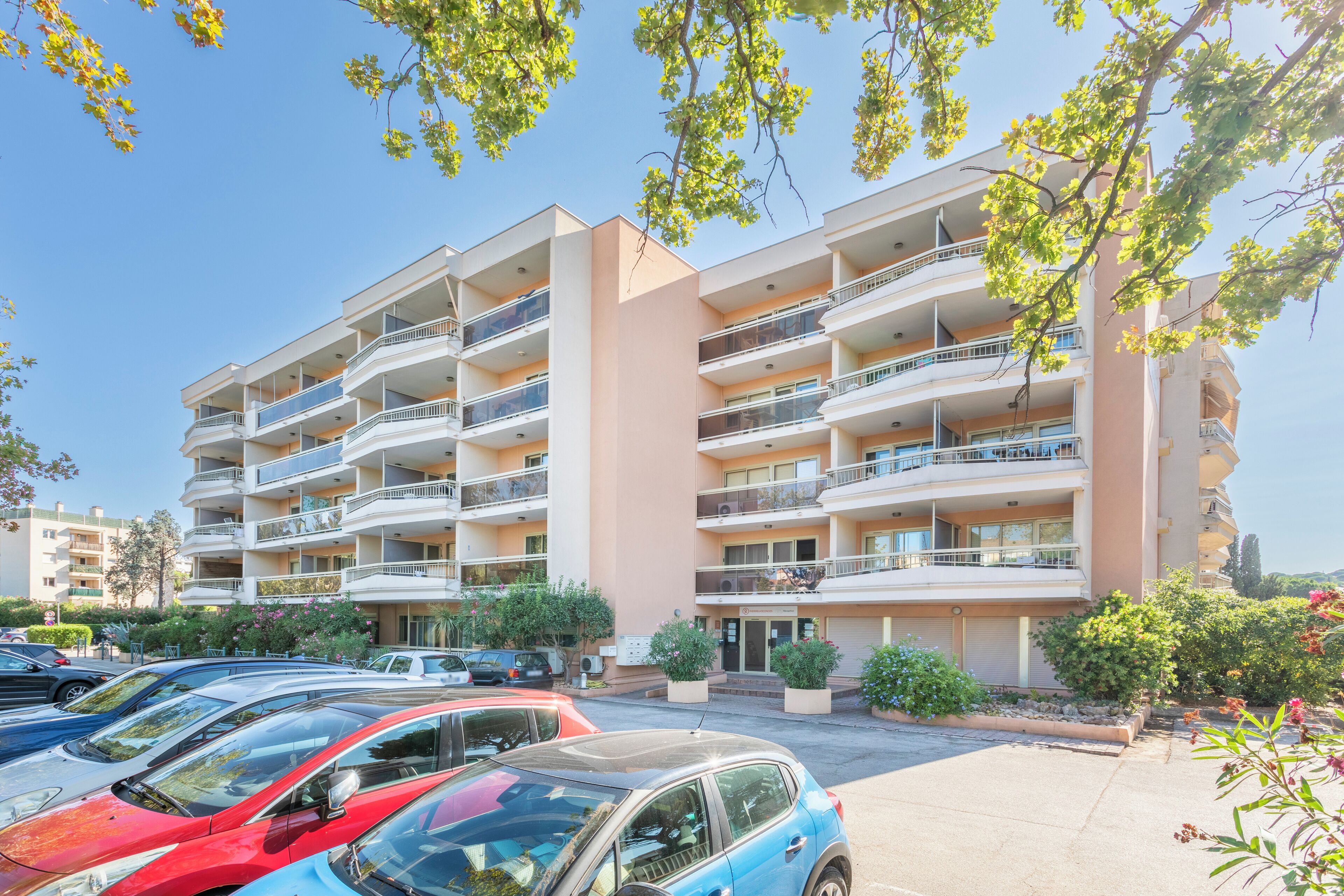 A multi-story apartment building with balconies, a parking lot, and surrounding greenery.