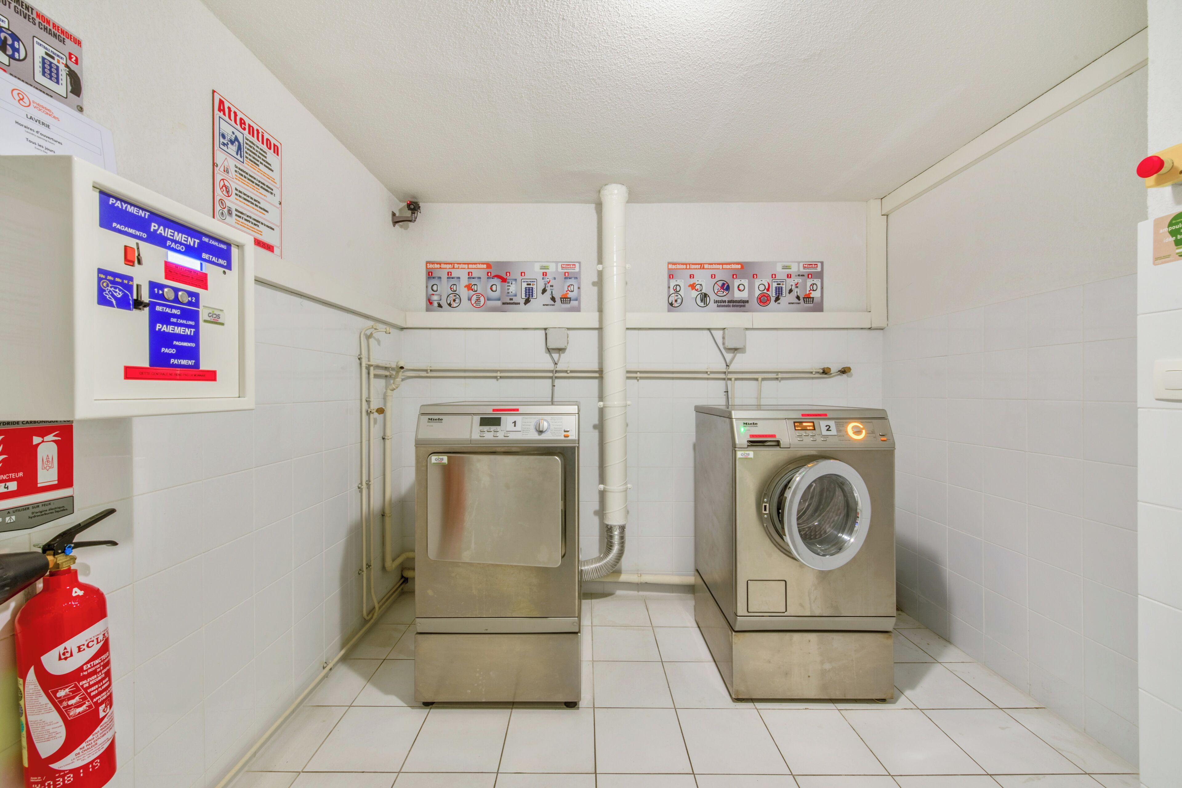 A laundry room with two stainless steel washers and dryers, a fire extinguisher, and safety signs on the walls.