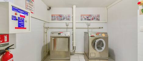 A laundry room with two stainless steel washers and dryers, a fire extinguisher, and safety signs on the walls.