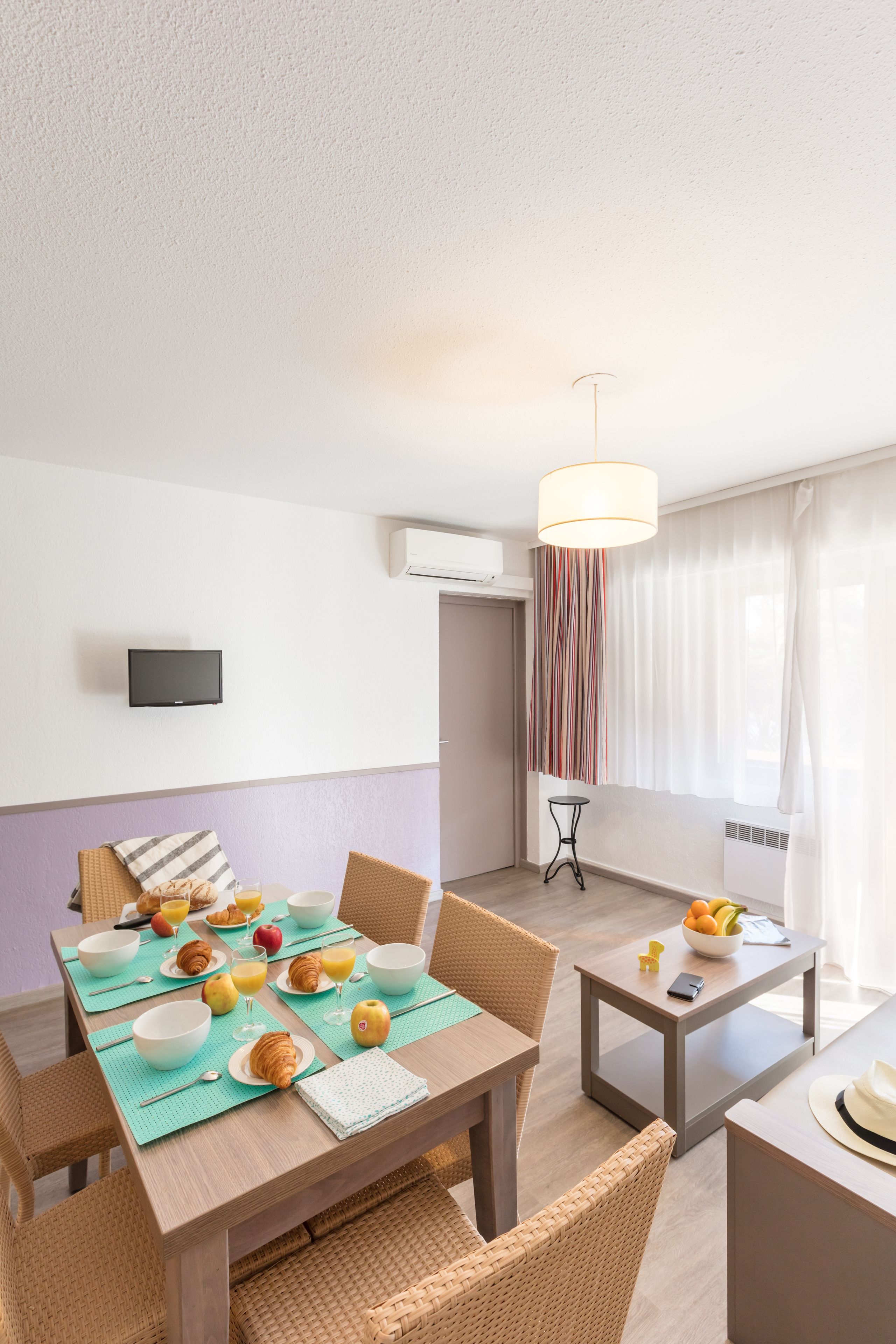 A dining area with a table set for breakfast, including croissants, fruit, and a bowl of oranges. There is a television on the wall, a hanging light fixture, and a window with curtains.