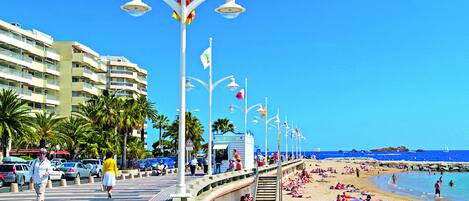A beachfront promenade with a row of beachside cafes, palm trees, and a clear blue sky.