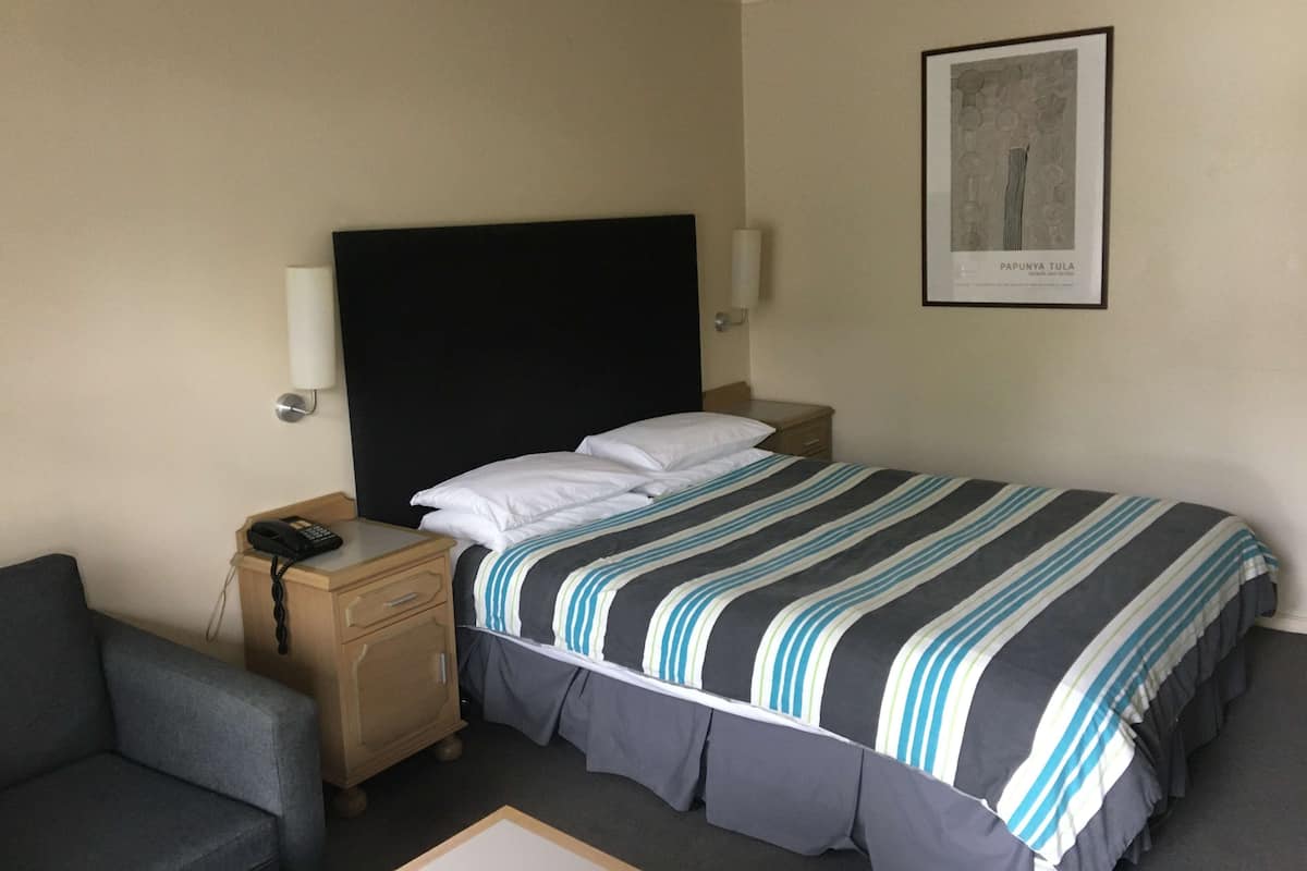 Ein Hotelzimmer mit Bett, Nachttischen, einem Sessel, einem gerahmten Bild an der Wand und einem Telefon auf dem Nachttisch.