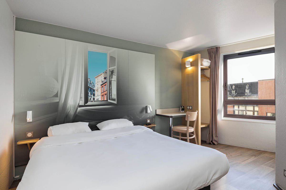 Quarto de hotel com uma cama grande, uma mesa, uma cadeira, uma janela com vista para prédios e um mural na parede com uma paisagem urbana.