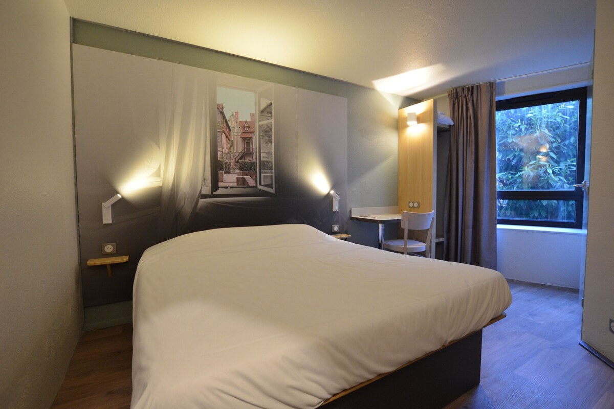 A hotel room with a bed, a desk, a chair, a window with curtains, and a wall-mounted lamp.