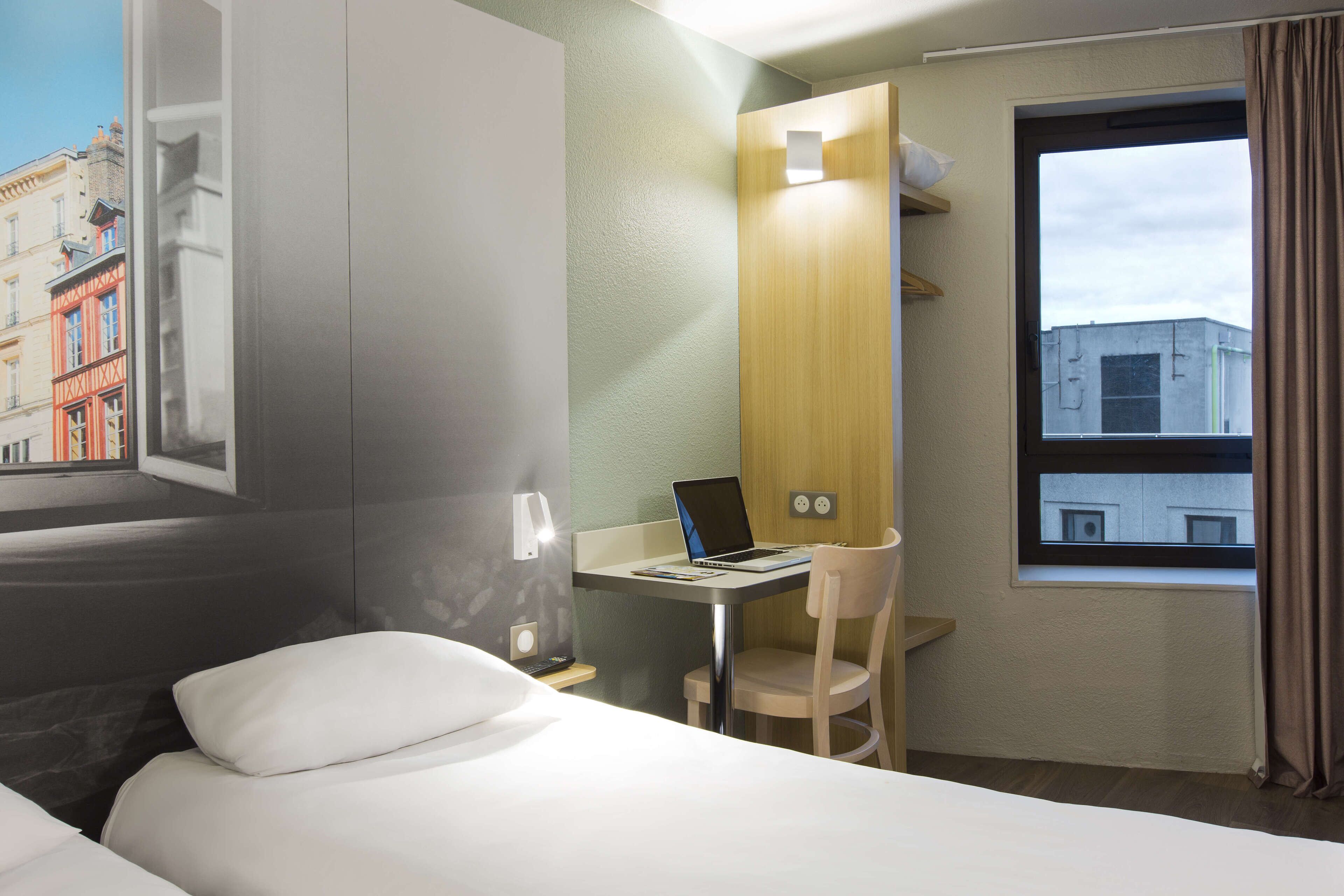 Habitación de hotel con cama, escritorio con una computadora portátil, silla y ventana con cortinas.