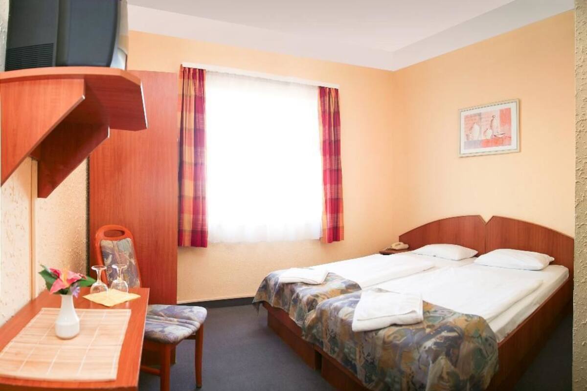 A hotel room with a bed, a desk with a chair, a vase with flowers, and a window with curtains.