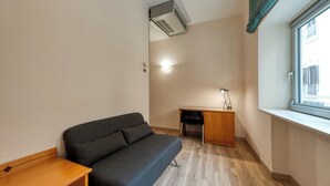 A small, modern room with a grey sofa, a wooden desk, and a window with a view of buildings. - Barsotti Hotel (Brindisi)