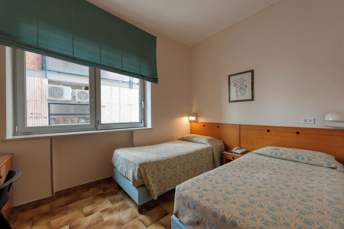 A hotel room with two beds, a window with blinds, a bedside table with a lamp, and a wall-mounted phone.