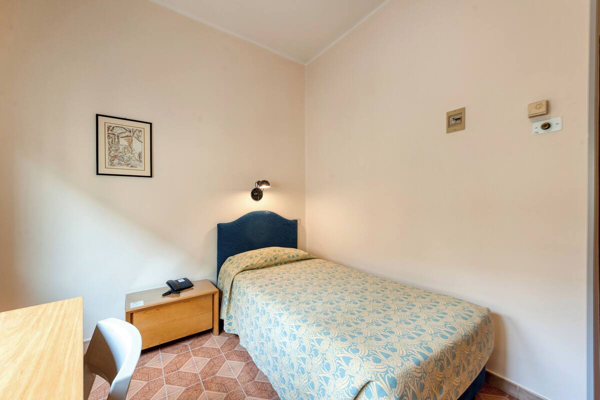 A bedroom with a single bed, a bedside table with a lamp, a chair, and a framed picture on the wall.
