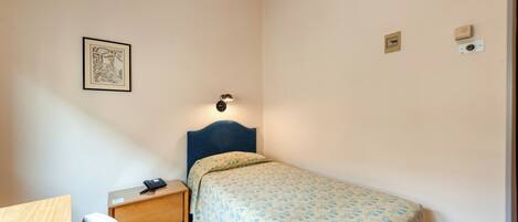 A bedroom with a single bed, a bedside table with a lamp, a chair, and a framed picture on the wall.