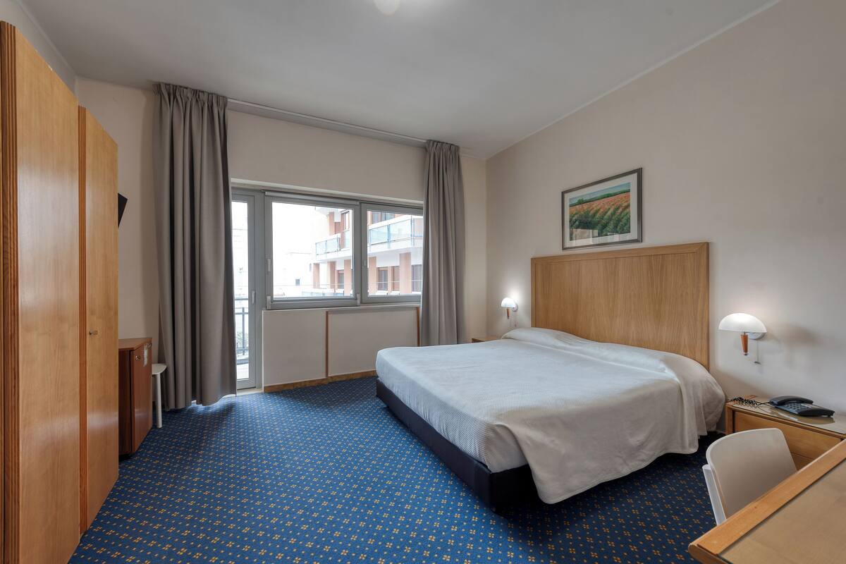 A hotel room with a large bed, a desk, a chair, a window with curtains, and a painting on the wall.